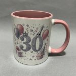 Close-up of a white birthday mug with pink interior and handle, decorated with balloons, stars, confetti, a crown, and the number 30.
