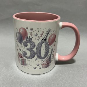 Close-up of a white birthday mug with pink interior and handle, decorated with balloons, stars, confetti, a crown, and the number 30.